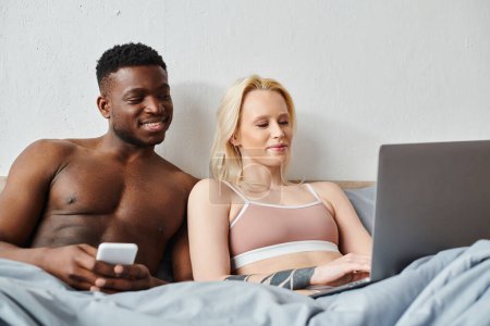 Multicultural couple sitting on bed, engrossed in laptop screen.