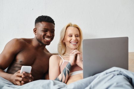 A multicultural couple sitting on a bed, engrossed in a laptop screen.