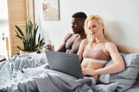 A multicultural man and woman are sitting on a bed, engrossed in the screen of a laptop in an intimate and cozy setting.