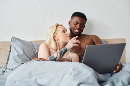 A multicultural couple relaxes in bed, engrossed in the content on their laptop screen.