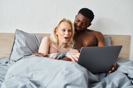 Foto de Una pareja multicultural mira la pantalla de un portátil en la cama. - Imagen libre de derechos