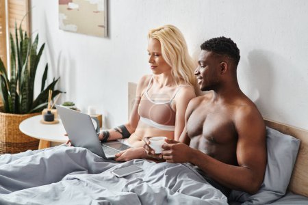 A multicultural couple in bed, immersed in a laptop screen.