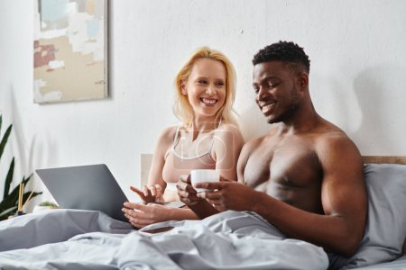 Un novio y una novia multiculturales se sientan en una cama, cautivados por la pantalla de un ordenador portátil.