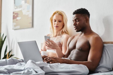 Foto de Un novio y una novia multiculturales se sientan en una cama, intensamente enfocados en la pantalla de un portátil. - Imagen libre de derechos