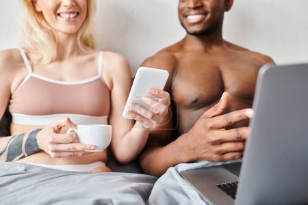 Un novio y una novia multiculturales sentados en una cama, centrándose en la pantalla de un ordenador portátil juntos.