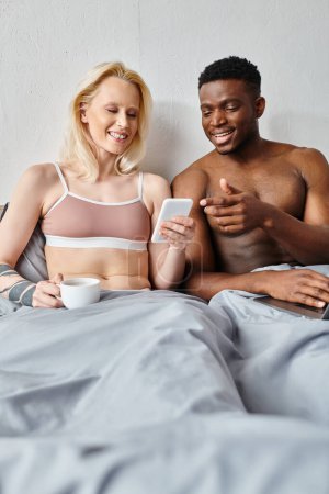 Téléchargez les photos : Un petit ami multiculturel et sa petite amie assis sur un lit, absorbés dans leur téléphone portable ensemble. - en image libre de droit