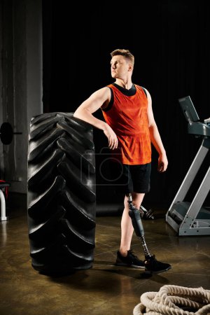 A disabled man with a prosthetic leg stands proudly next to a large tire in a gym, showcasing his determination and strength.