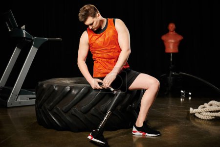 A man with a prosthetic leg sits on a tire next to a machine, deep in thought.