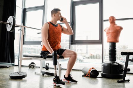 A man with a prosthetic leg sits in gym, engaged in a phone conversation amidst urban surroundings.