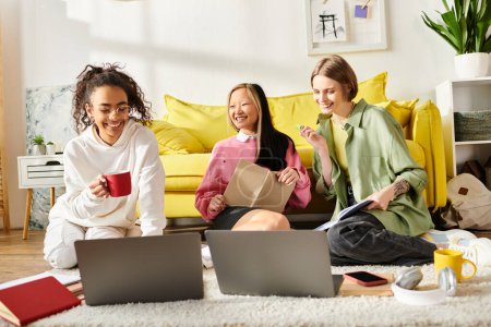 Tres adolescentes de diferentes razas se sientan en el suelo con computadoras portátiles, absortas en sus estudios y fomentando un vínculo de amistad a través de la educación.