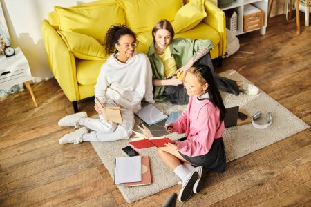 Three interracial teenage girls sit on the floor in front of a yellow couch, studying together in the comfort of their home.