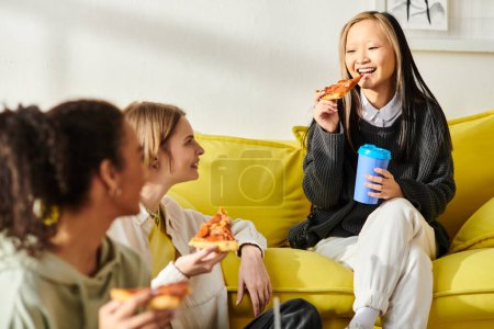 Three teenage girls of different races sit on a yellow couch, enjoying pizza together.