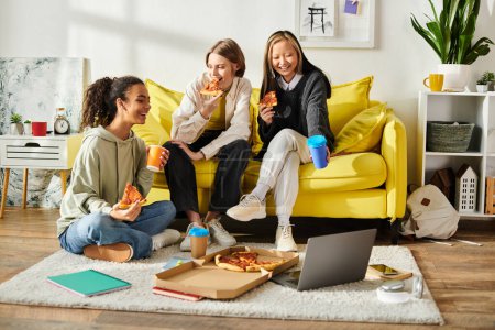 Three teenage girls of diverse backgrounds laugh and chat while sitting on a yellow couch, enjoying slices of pizza together.