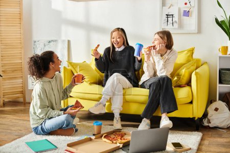 Three teenage girls, of different ethnicities, are seated on a bright yellow couch, enjoying slices of pizza together.