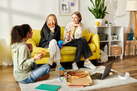 Trois jeunes femmes de races différentes dégustant une pizza assis sur un canapé jaune vibrant.