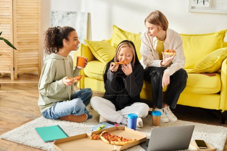 Three young women of different races and styles sit on the floor, enjoying pizza together in a cozy setting.