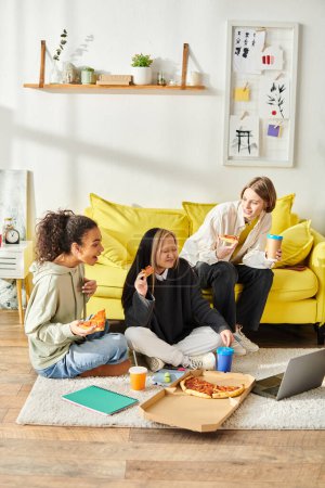 Eine bunte Gruppe Teenager-Mädchen sitzt auf dem Fußboden und isst zu Hause fröhlich gemeinsam Pizza..