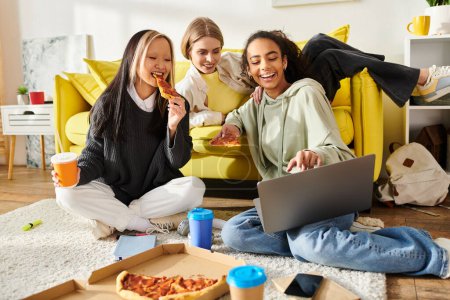 Three teenage girls of different ethnicities sit on the floor, enjoying pizza and using a laptop together.