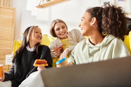 Eine bunte Gruppe Teenager-Mädchen lacht und plaudert, während sie auf einer Couch sitzen und gemeinsam leckere Pizza essen.