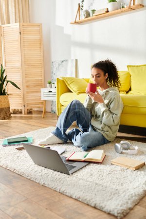A young woman sits on the floor, savoring a cup of coffee while engaged in e-learning on her laptop.