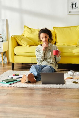 Eine junge Frau sitzt auf dem Boden und klammert sich anmutig an eine warme Tasse Kaffee, während sie zu Hause an ihrem Laptop lernt..