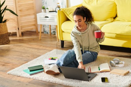 Téléchargez les photos : Une adolescente assise sur le sol, absorbée par l'apprentissage en ligne sur son ordinateur portable, accompagnée d'une tasse de café. - en image libre de droit