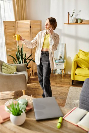 Photo for A teenage girl stands in a living room, talking on a cell phone while e-learning with a laptop. - Royalty Free Image