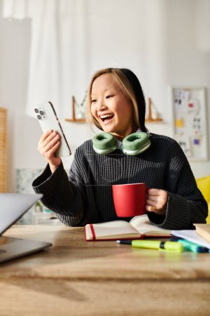 Una joven asiática se sienta en una mesa, con un portátil abierto, con una taza de café frente a ella.
