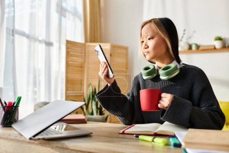 Téléchargez les photos : Une jeune femme asiatique étudie à la maison, sirotant paisiblement un café assis à une table avec un ordinateur portable. - en image libre de droit