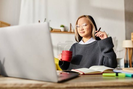 A young Asian woman sits at a table, studying at home with a laptop and a cup of coffee.