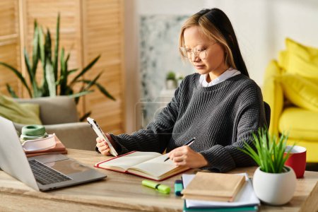A young Asian girl sitting at a desk, deeply focused on a laptop and notebook while studying at home.