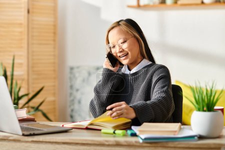 Photo for A young Asian woman sitting at a desk, engaged in a phone call while studying with a laptop for e-learning. - Royalty Free Image