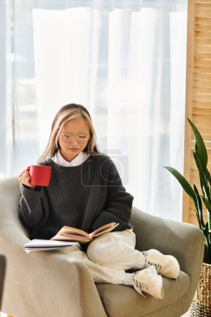 A young Asian woman sits in a chair, engrossed in a book while sipping coffee.