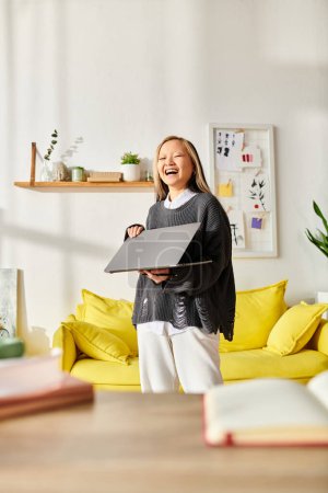 Ein junges asiatisches Mädchen steht in einem Wohnzimmer und beschäftigt sich intensiv mit E-Learning, hält einen Laptop in der Hand.