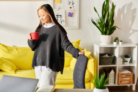 Una joven asiática de pie en una sala de estar, disfrutando de una taza de café mientras aprende en línea con su computadora portátil.