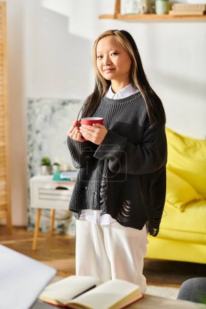 A young Asian woman stands inside a cozy living room, joyfully holding a cup as she takes a satisfying sip.