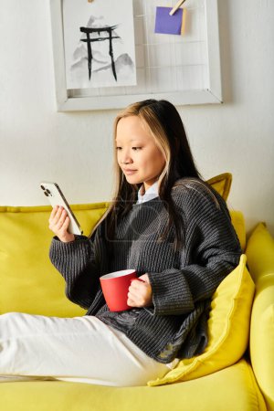 A young Asian woman sits on a cozy yellow couch, holding a cup of coffee.