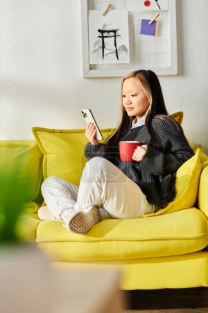 A young Asian woman sits on a yellow couch, holding a cup of coffee while studying on her smartphone in a cozy home setting.