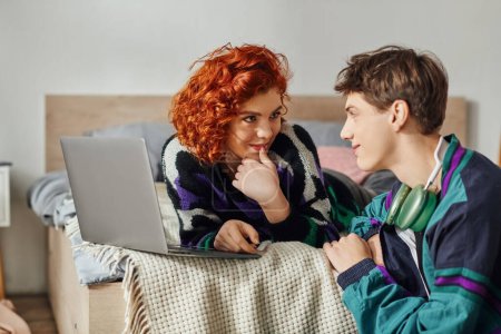 Téléchargez les photos : Femme aux cheveux rouges en tenue confortable assis sur le lit et passer du temps avec son petit ami joyeux à l'ordinateur portable - en image libre de droit