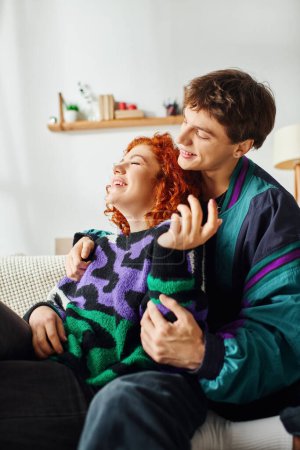 Téléchargez les photos : Beau couple gai dans des vêtements confortables à la mode se serrant les uns les autres tandis qu'à la maison près du lit - en image libre de droit