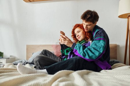 Téléchargez les photos : Homme attrayant avec écouteurs étreignant sa petite amie gaie aux cheveux roux alors qu'il était au lit à la maison - en image libre de droit