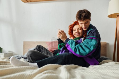 Téléchargez les photos : Joyeux bel homme avec écouteurs étreignant sa petite amie avec amour alors qu'il était à la maison sur le lit - en image libre de droit