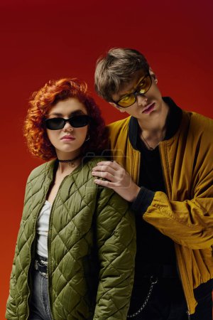 Téléchargez les photos : Attrayant couple à la mode avec des lunettes de soleil à la mode dans des vêtements confortables posant sur fond rouge ensemble - en image libre de droit