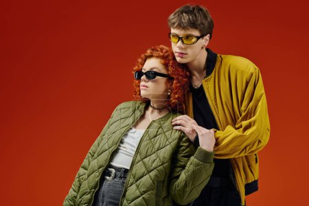 Téléchargez les photos : Séduisant couple à la mode avec des lunettes de soleil fraîches dans des vêtements confortables posant sur fond rouge ensemble - en image libre de droit