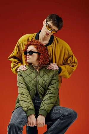 handsome man posing next to his red haired girlfriend who sitting on chair on red background