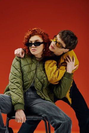 appealing red haired woman sitting on chair next to her stylish boyfriend, both in sunglasses