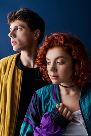 Téléchargez les photos : Séduisante petit ami et petite amie aux cheveux rouges posant ensemble sur fond bleu foncé et regardant loin - en image libre de droit