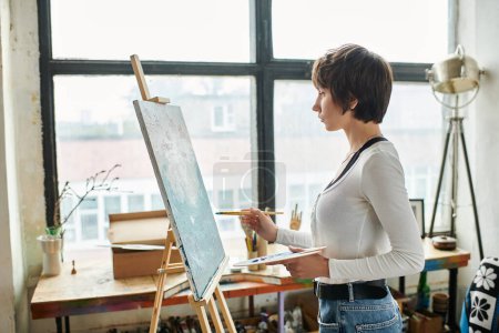 A woman stands confidently in front of a painting easel in an art studio.