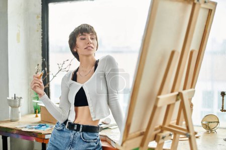 Woman standing, holding paintbrush in front of easel.
