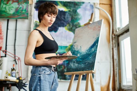 Foto de Una mujer en una camiseta negra se centra en la pintura en un caballete. - Imagen libre de derechos
